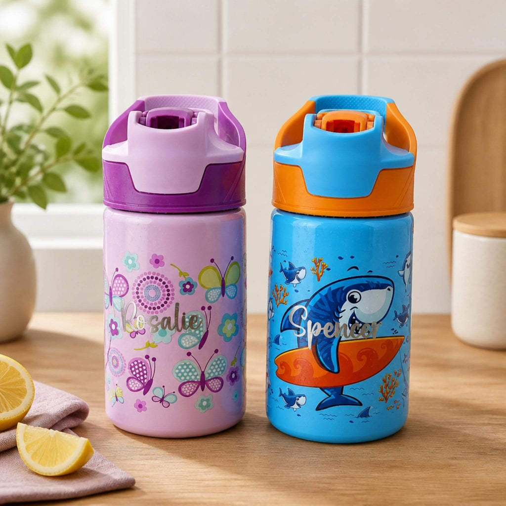 Two personalised kids water bottles on a wooden benchtop, one purple with a butterfly and floral design and the name "Rosalie", one blue with a surfing shark design and the name "Spencer" — Little Cutlery Australia