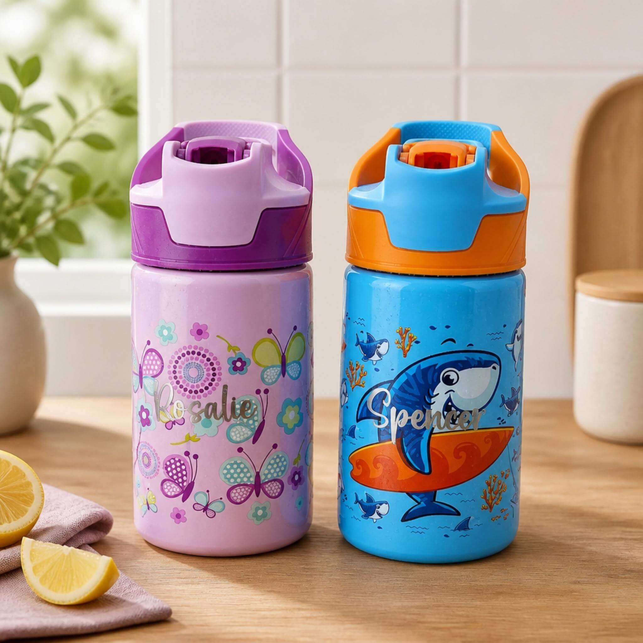 Two personalised kids water bottles on a wooden benchtop, one purple with a butterfly and floral design and the name "Rosalie", one blue with a surfing shark design and the name "Spencer" — Little Cutlery Australia