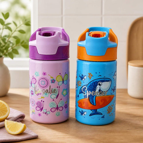 Two personalised kids water bottles on a wooden benchtop, one purple with a butterfly and floral design and the name "Rosalie", one blue with a surfing shark design and the name "Spencer" — Little Cutlery Australia