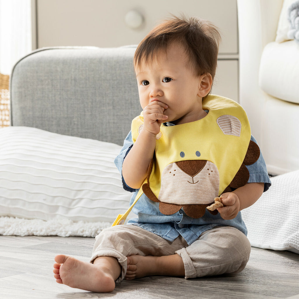 Toddler sitting on the floor wearing a yellow cotton bib with an applique lion design while eating a snack