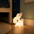 Glowing bunny-shaped silicone LED night light sitting on the floor beside a bed in a dimly lit children's bedroom
