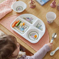 Toddler seated at a table with a dinosaur print divided plate of vegetables, matching bowl and cup, on a pink placemat
