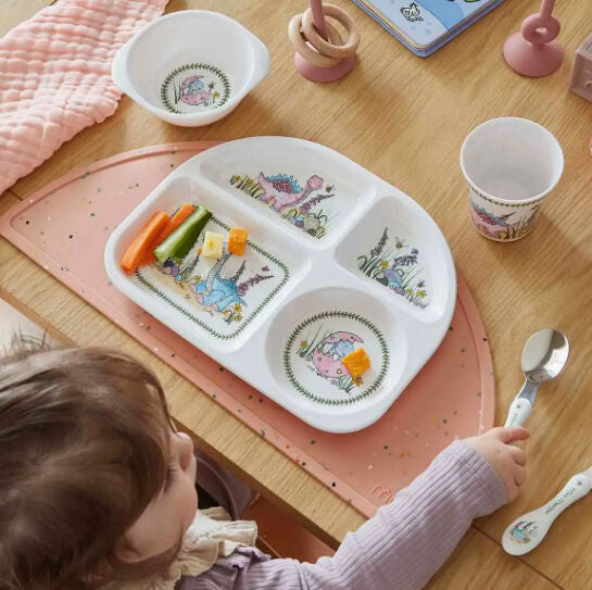 Toddler seated at a table with a dinosaur print divided plate of vegetables, matching bowl and cup, on a pink placemat