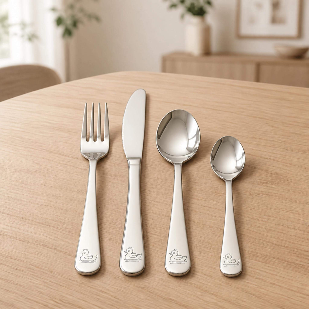 Four-piece stainless steel children's cutlery set with a laser-engraved duck design on each handle, laid out on a wooden table