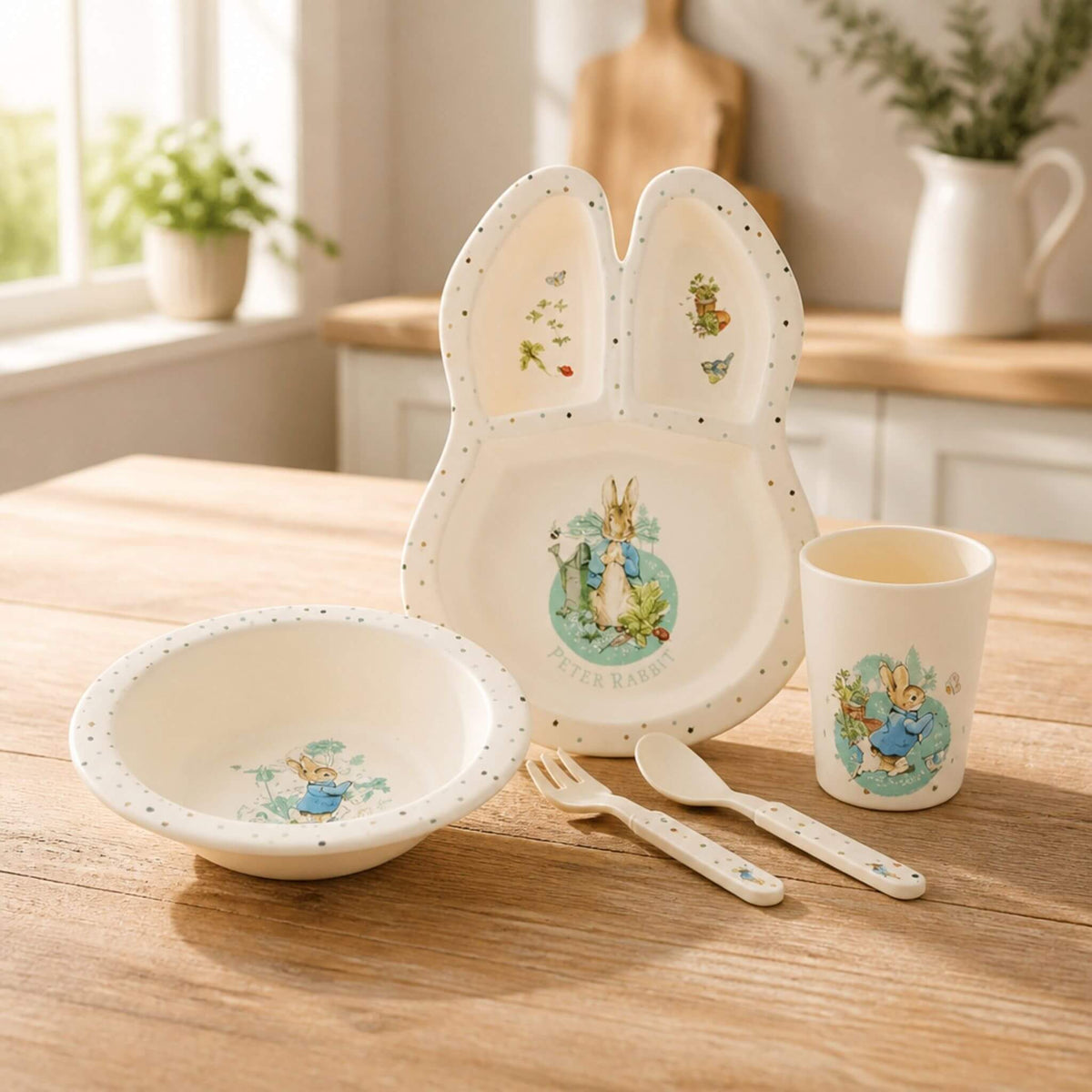 Peter Rabbit dinnerware set featuring a bunny-ear shaped divided plate, bowl, cup, fork and spoon on a wooden kitchen table