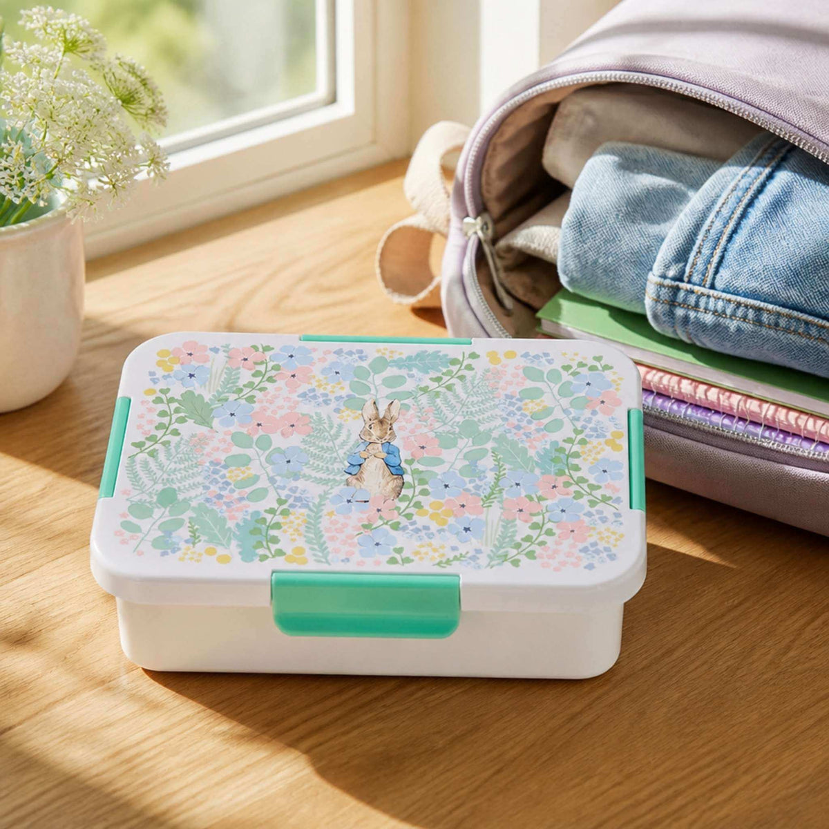 Peter Rabbit floral print bento lunch box with mint green clasps on a windowsill beside a backpack, notebooks and flowers