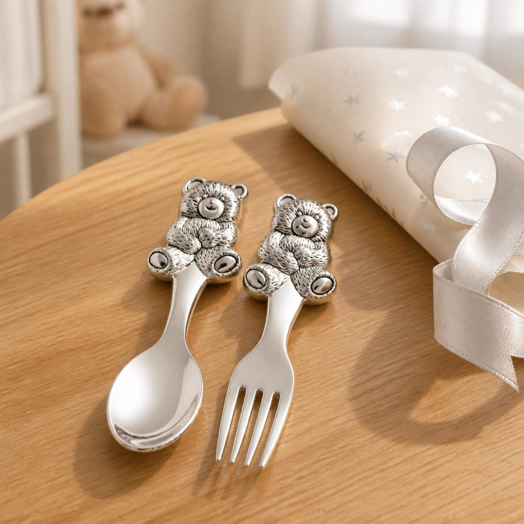 Silver-plated teddy bear spoon and fork set displayed on a wooden table with a nursery cushion, ribbon and teddy bear in the background