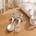 Silver-plated teddy bear spoon and fork set displayed on a wooden table with a nursery cushion, ribbon and teddy bear in the background